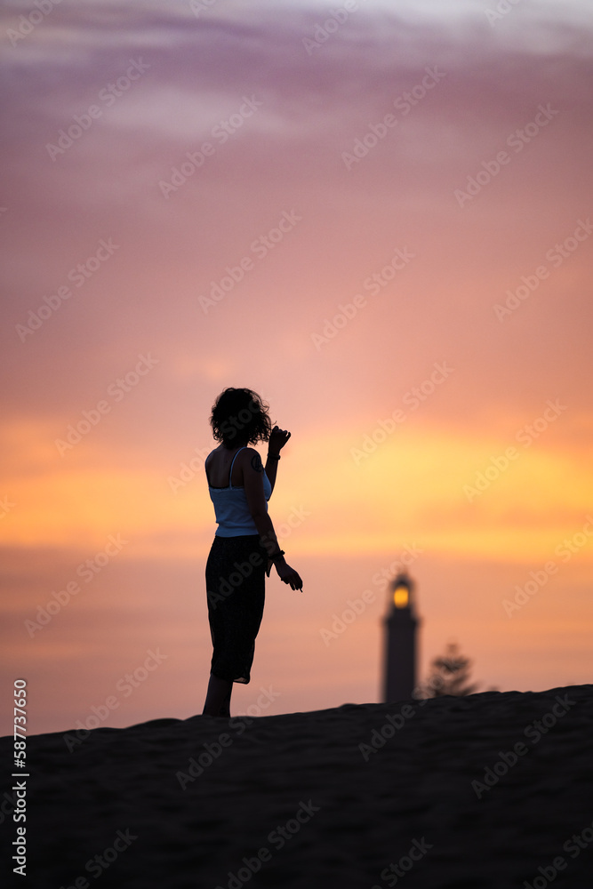 Stunning view of a silhouette of a person walking on some sand dunes at sunset with the Maspalomas lighthouse in the distance. Gran Canaria, Canary Islands