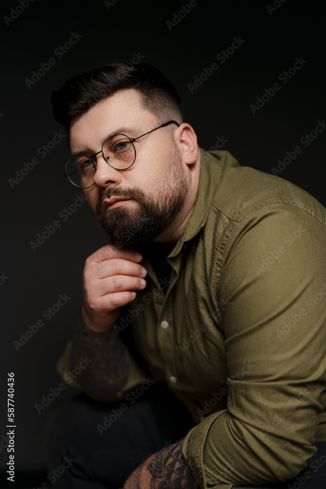 Thoughtful young plump tattooed male model in eyeglasses and green shirt touching beard against black background in studio Thoughtful young plump tattooed male model in eyeglasses and green shirt touching beard against black background in studio