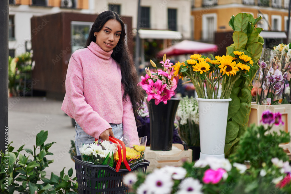 A young Latina woman shopping for plants, carrying a basket full of them in a street florist shop.