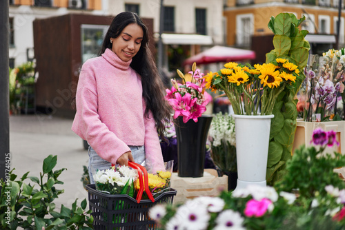 A young Latina woman shopping for plants, carrying a basket full of them in a street florist shop.