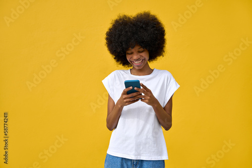 A hipster girl with afro haircut enjoy online communication and online services, wear trendy T-shirt, scroll social media apps on cell phone device isolated on yellow wall. Technology and applications
