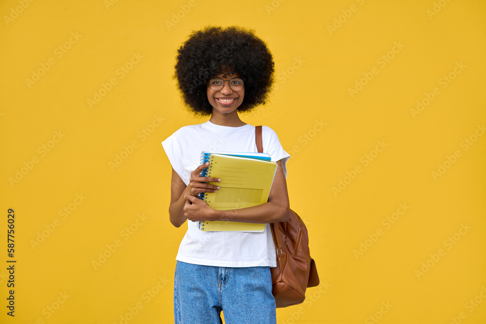 Young happy african american girl with curly afro hairstyle stands with backpack and holds exercise books on a yellow background. Smart student looking at camera on orange wall with copy space. Young happy african american girl with curly afro hairstyle stands with backpack and holds exercise books on a yellow background. Smart student looking at camera on orange wall with copy space.