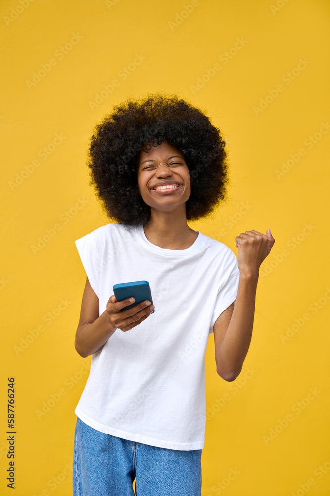 A vertical portrait of happy hipster student girl with afro haircut showing victory triumph gesture when using social media app on cell phone device isolated on yellow wall. Technology and application A vertical portrait of happy hipster student girl with afro haircut showing victory triumph gesture when using social media app on cell phone device isolated on yellow wall. Technology and application