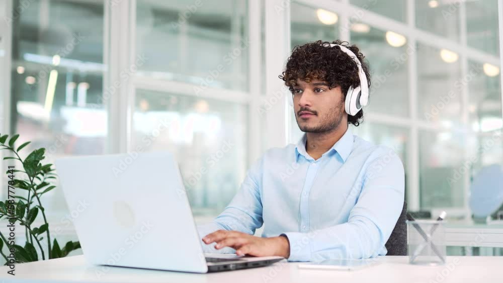 Young smiling worker in headphones typing on laptop while sitting at workplace at desk in modern office. A handsome developer in a shirt works on a project while listening to his favorite music Young smiling worker in headphones typing on laptop while sitting at workplace at desk in modern office. A handsome developer in a shirt works on a project while listening to his favorite music