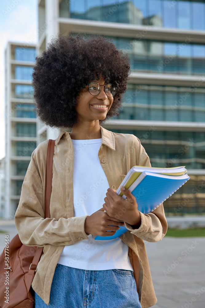A vertical portrait of African American girl with curly afro hairstyle stands with backpack and holds exercise books in university campus. Smart student holding books looking aside on city background. A vertical portrait of African American girl with curly afro hairstyle stands with backpack and holds exercise books in university campus. Smart student holding books looking aside on city background.