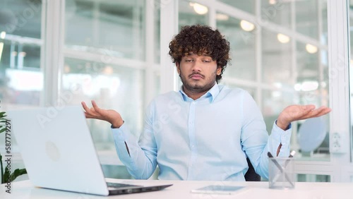 Young surprised puzzled worker spreads hands, shrugs shoulders looking at camera working on laptop while sitting at workplace at desk in office. Man in the shirt has a confused expression on his face