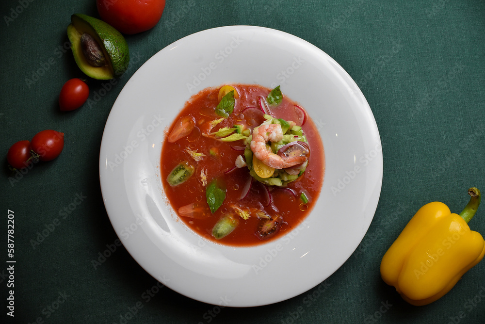 Shrimp with cherry tomatoes, avocado, cucumbers, and radishes in a red sauce on a white plate against a dark green background. Shrimp with cherry tomatoes, avocado, cucumbers, and radishes in a red sauce on a white plate against a dark green background.