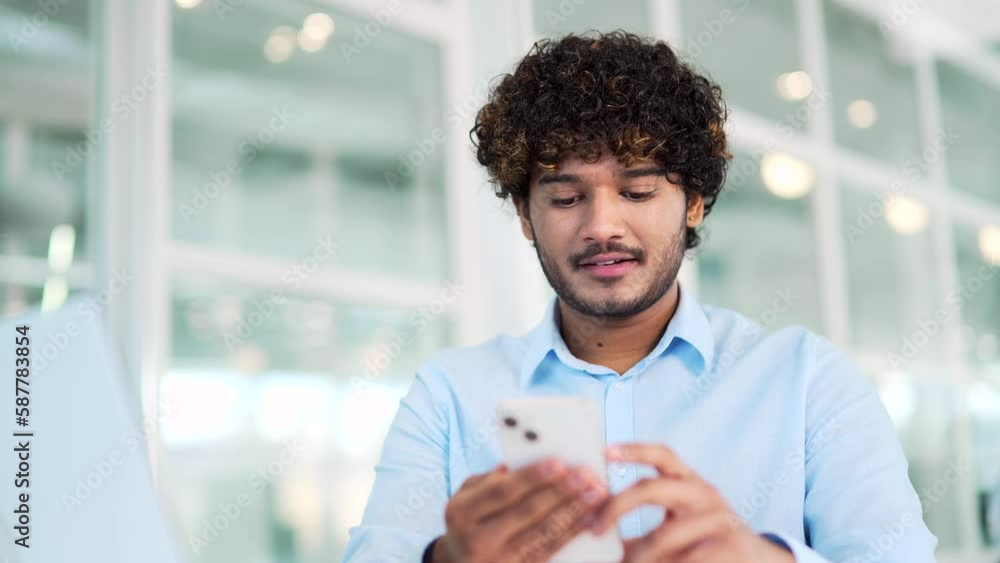 Close up. A young handsome worker is using a smartphone while sitting at a workplace at a desk in a office. A smiling employee is browsing social networks, chatting online, typing or reading a message Close up. A young handsome worker is using a smartphone while sitting at a workplace at a desk in a office. A smiling employee is browsing social networks, chatting online, typing or reading a message