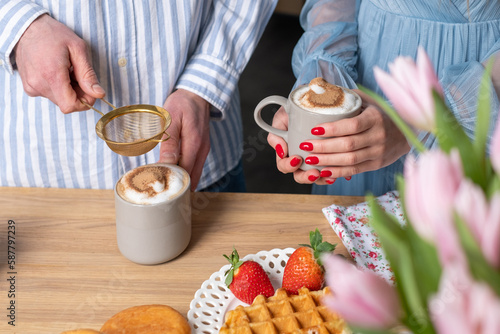 A mans and a woman s hands in light blue clothes with red nails. A man ads cinnamon in a gray cup with caffe latte. The crispy waffle  red strawberries on a white plate and donuts.