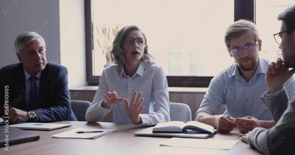 Middle-aged businesswoman makes speech during formal meeting, negotiate with corporate clients seated at desk in modern conference room. Female boss instruct team, explain tasks, provide information Middle-aged businesswoman makes speech during formal meeting, negotiate with corporate clients seated at desk in modern conference room. Female boss instruct team, explain tasks, provide information