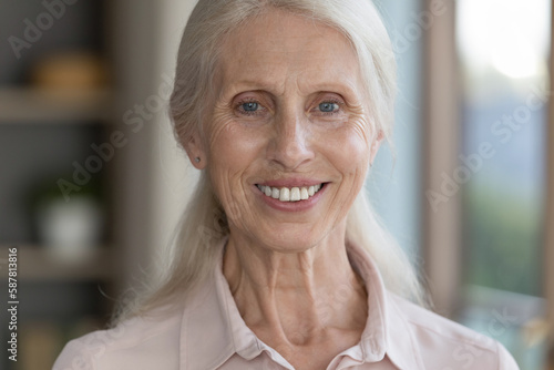 Happy pretty old senior lady looking at camera with toothy smile, posing at home. Elderly blonde grey haired woman, beauty care, healthcare model head shot portrait