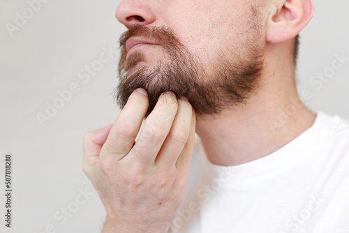 Cropped shot mans hand is unrestrained scratching unshaven chin. Overgrown unkempt male in white t shirt with thick beard and mustache face. Beauty salon concept, barbershop