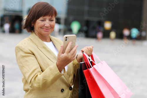 Stylish adult woman blogger with shopping bags and smartphone is communicating with followers in social networks near shopping mall. concept of consumerism, sale, rich life, virtual life. Black Friday