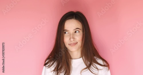 Playful happy flirt brunette woman coquettish look by side, blinking eye, looking at camera with smile, winking and flirting, expressing optimism isolated on pink background.