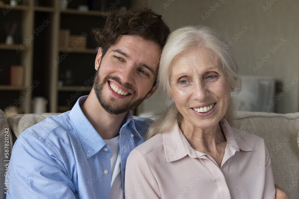 Happy grandson guy and positive older grandma sitting close on home sofa, hugging, looking away with toothy smiles. Family portrait of handsome adult son and senior mom Happy grandson guy and positive older grandma sitting close on home sofa, hugging, looking away with toothy smiles. Family portrait of handsome adult son and senior mom