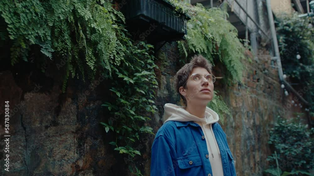 Young short-haired girl in casual outfit and eyeglasses standing by a brick wall, looking at fern growing on it, visiting the old town. Waist-up shot, copy space Young short-haired girl in casual outfit and eyeglasses standing by a brick wall, looking at fern growing on it, visiting the old town. Waist-up shot, copy space