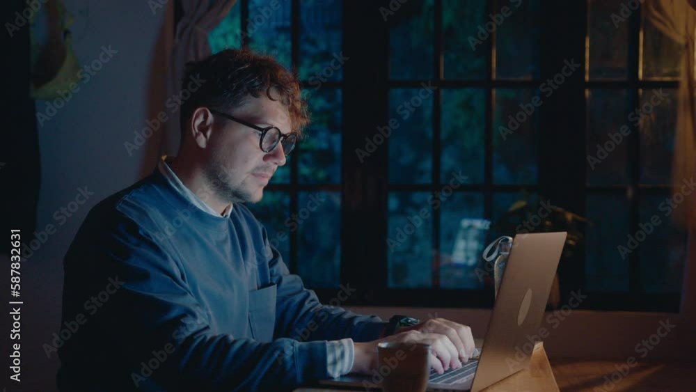 Young man in eyeglasses typing on laptop in dark room, working remotely overtime during night. Medium shot Young man in eyeglasses typing on laptop in dark room, working remotely overtime during night. Medium shot