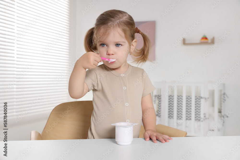 Cute little child eating tasty yogurt from plastic cup with spoon at white table indoors Cute little child eating tasty yogurt from plastic cup with spoon at white table indoors