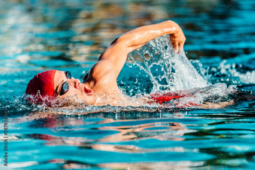 Male swimmer swimming crawl freestyle in blue water outdoor pool. Portrait of an athletic young male triathlete swimming crawl wearing a red cap and swim goggles. Triathlete training for triathlon