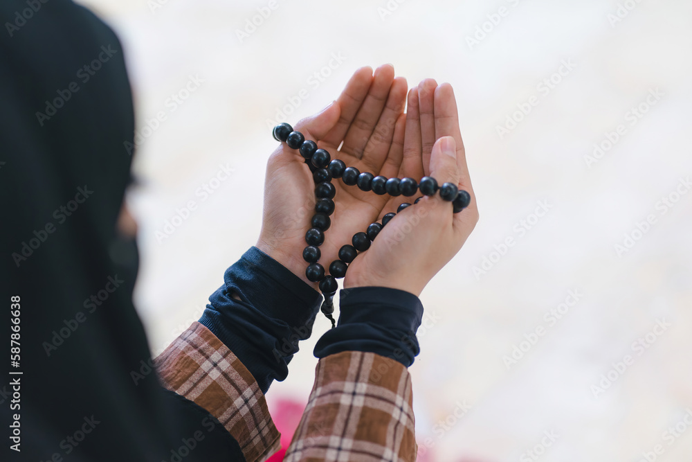 A close-up shot hands of a Muslim woman holding a rosary while wearing a long hijab, as she prays and makes dua to Allah, the Muslim God, at the mosque during an Islamic religious ceremony.