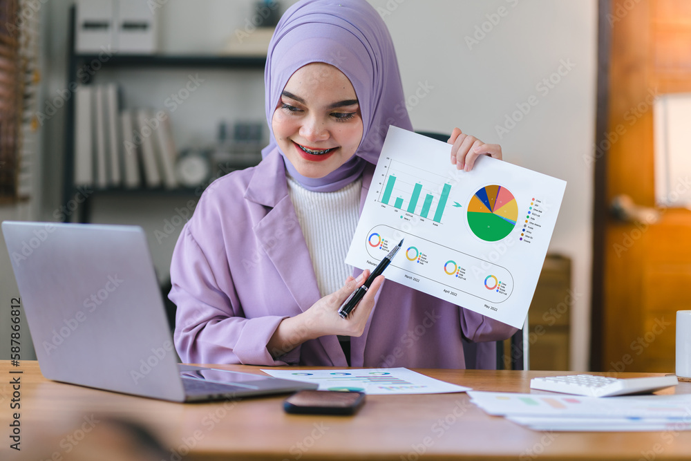 A Muslim millennial businesswoman, wearing a purple hijab, is shown working with financial report paperwork in home office. The concept of financial advising, teamwork, and accounting is depicted. A Muslim millennial businesswoman, wearing a purple hijab, is shown working with financial report paperwork in home office. The concept of financial advising, teamwork, and accounting is depicted.