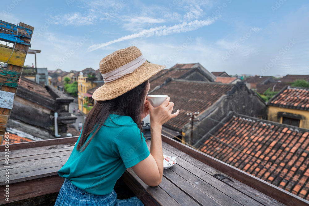 Young tourist woman in a hat sits on the roof of a coffee shop drinking coffee overlooking the old town of Hoi An Early morning in Da Nang city, Vietnam