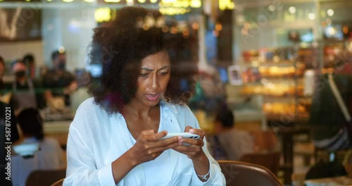 Excited emotional and worried young African American woman, sitting in a cafeteria, focused on playing computer game on her smartphone, expressing anger and negative emotions while failing the round photo