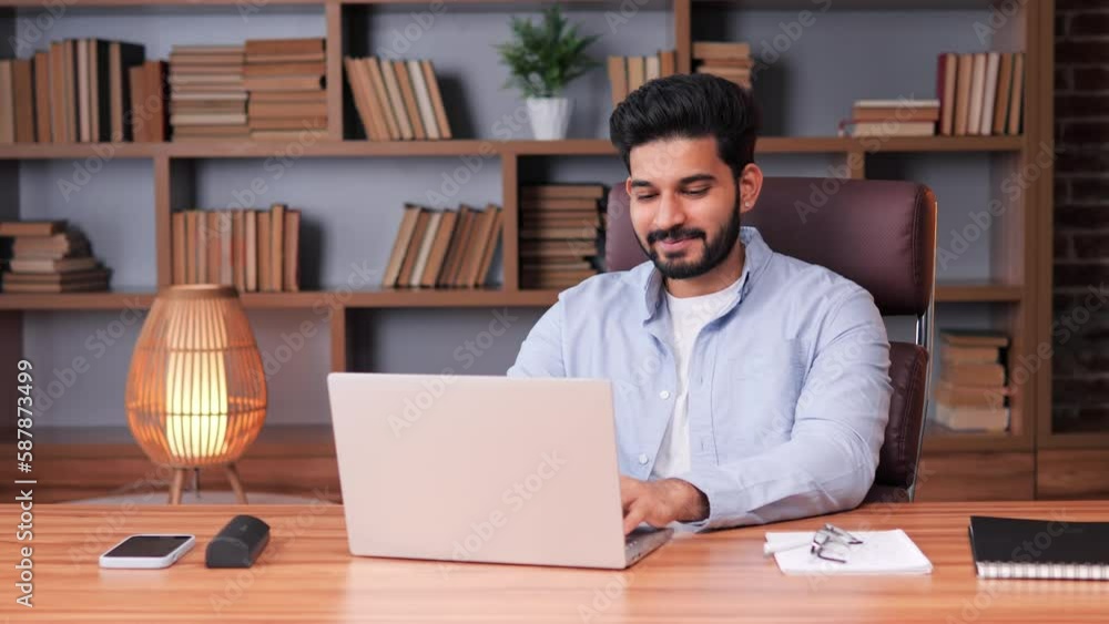 An upbeat, cheerful Indian businessman in a blue shirt sitting in office, using laptop to communicate. Business man looking ahead and appears lost in thought. Man sending message in social media. An upbeat, cheerful Indian businessman in a blue shirt sitting in office, using laptop to communicate. Business man looking ahead and appears lost in thought. Man sending message in social media.