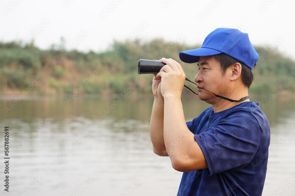 Asian man wears blue cap, hold binoculus to explore something at lake, nature source.Concept, nature exploration. Ecology study.  Pastime activity, lifestyle. Man explore environment          