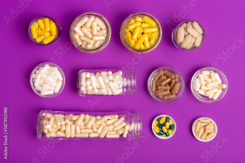 Many small glass bottles and bowls filled with different vitamins and minerals in form of capsules, tablets and pills from above on violet background. Vitamin b, d3, c, a, e, magnesium, zinc.