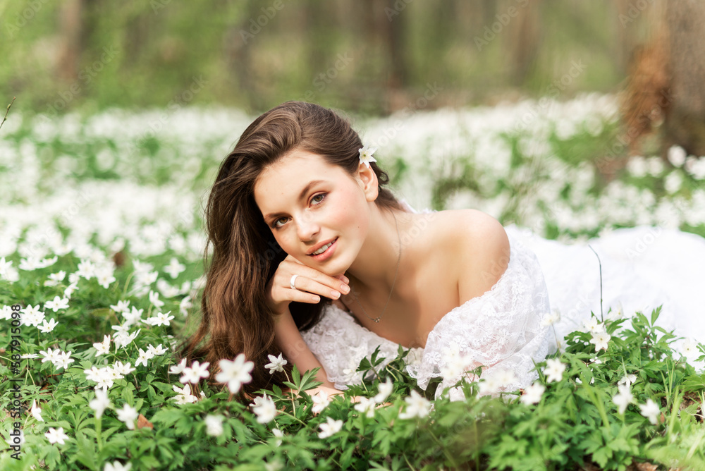 Portrait of a beautiful girl with long hair in a spring forest. A girl in a white lace dress lies in a clearing with primroses Portrait of a beautiful girl with long hair in a spring forest. A girl in a white lace dress lies in a clearing with primroses