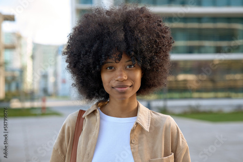 Closeup of emotional African American young woman girl with curly afro hairstyle in eyeglasses stands outside on city streets. Smart serious student looking at camera on blurry buildings background.