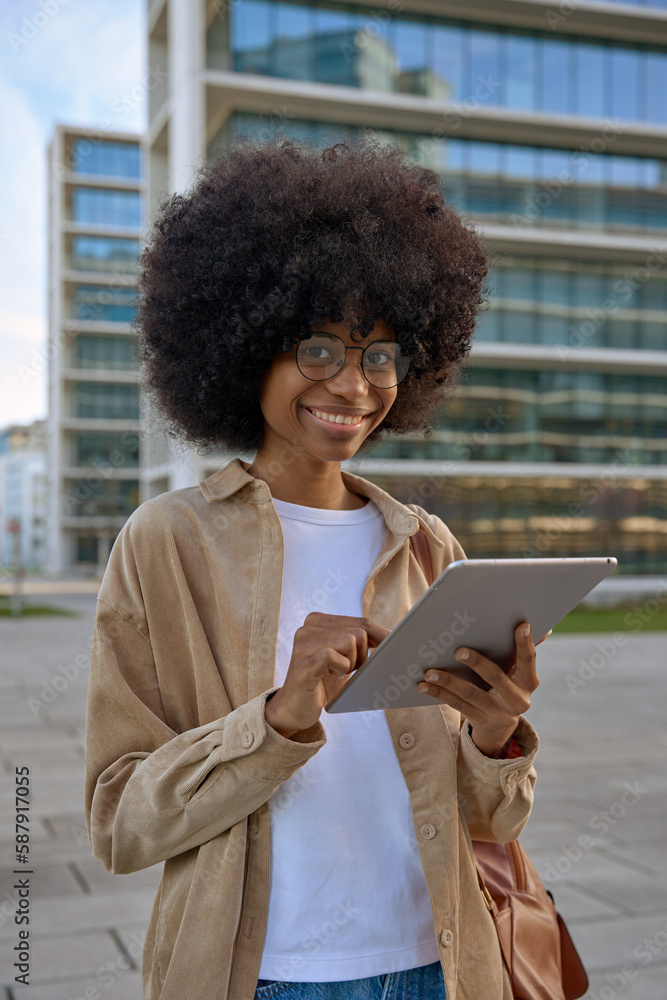 Vertical portrait of young woman student using touchscreen of pc tablet standing in university campus. An African American female business freelancer entrepreneur using app for online work outdoors. Vertical portrait of young woman student using touchscreen of pc tablet standing in university campus. An African American female business freelancer entrepreneur using app for online work outdoors.