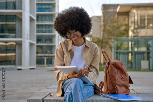 Happy young black African American woman university student doing homework, learning and taking notes in notepad sitting outside uni campus area on fresh air, near trees and buildings and smiling.