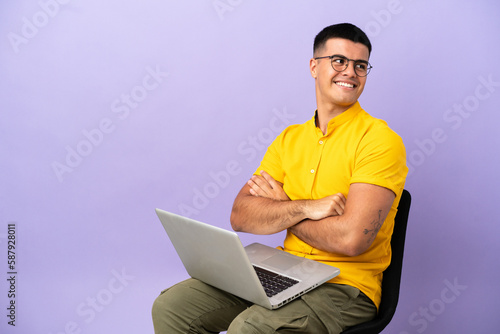 Young man sitting on a chair with laptop with arms crossed and happy