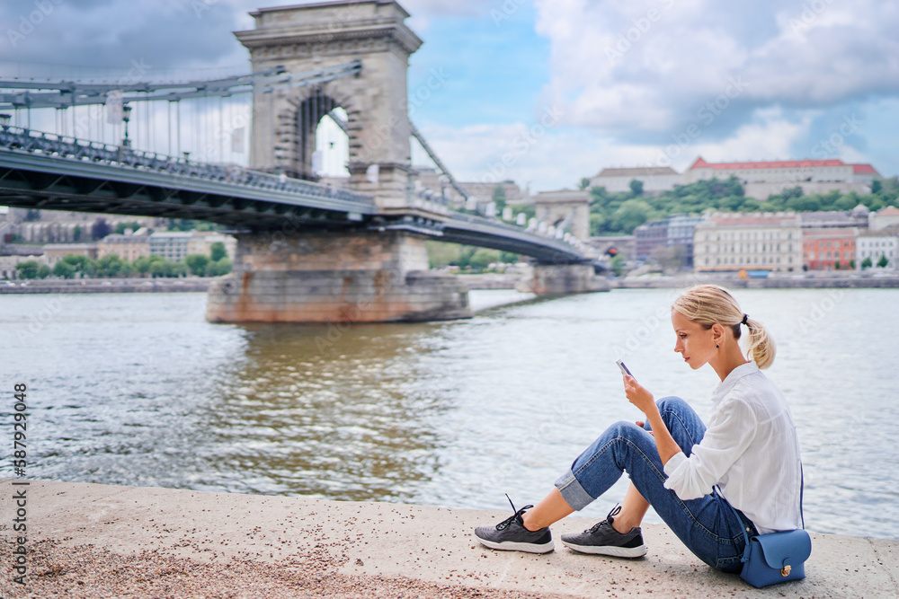 Vacation in Budapest. Young traveling woman using smartphone on riverside promenade with city view.