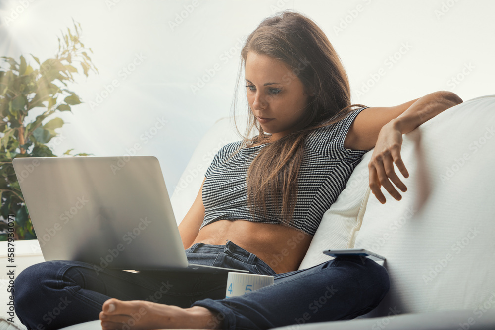 Focused woman on white couch, using grey laptop and enjoying tea Focused woman on white couch, using grey laptop and enjoying tea