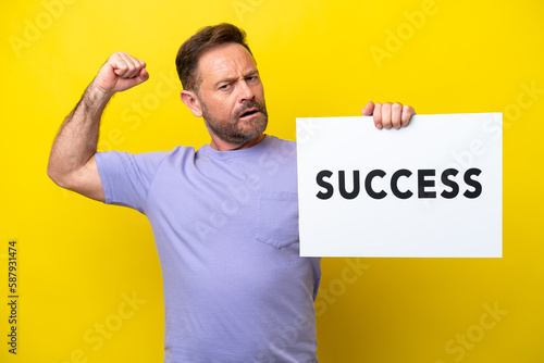 Middle age caucasian man isolated on yellow background holding a placard with text SUCCESS and doing strong gesture © luismolinero