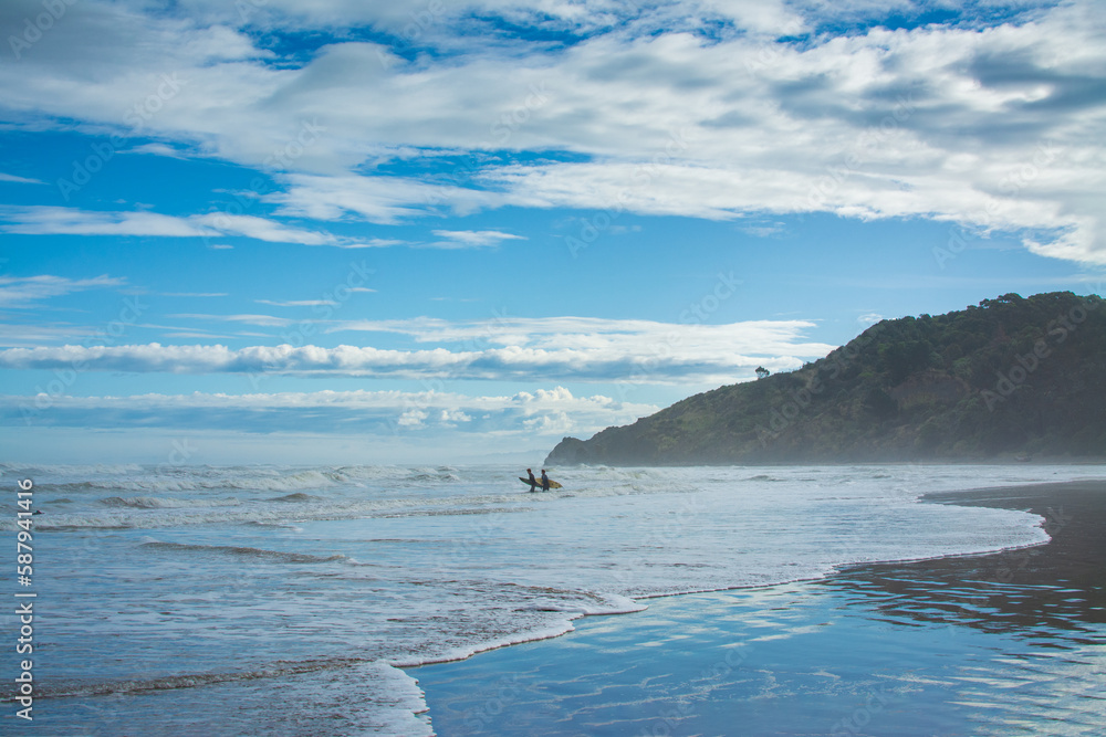 A pair of surfers walking into breaker water at Makorori Beach, mist rising over water obscuring distant mountains. Gisborne, New Zealand