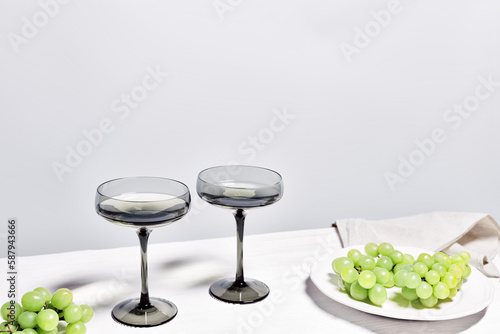 Dark shampagne glasses and grape on white wooden table, wine glass colored. Minimal aesthetic still life with sparkling white wine in wide shampagne glass, alcohol drink. copy space photo