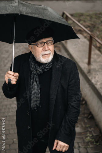 An elderly man in a black coat, glasses with an umbrella stands on the stairs in the rain, rainy weather