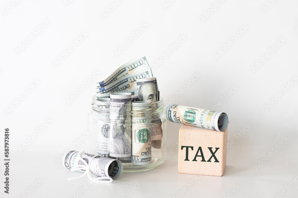 Rolls of savings and TAX inscription on a wooden cube. Payment and tax concept. Rolls of dollar in a jar with TAX writing Rolls of savings and TAX inscription on a wooden cube. Payment and tax concept. Rolls of dollar in a jar with TAX writing