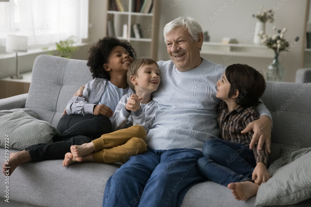 Cheerful old retired granddad hugging, cuddling cute little multiethnic grandsons, enjoying family meeting with boys, chatting, talking to grandchildren, smiling, laughing,