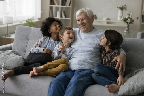 Cheerful old retired granddad hugging, cuddling cute little multiethnic grandsons, enjoying family meeting with boys, chatting, talking to grandchildren, smiling, laughing,