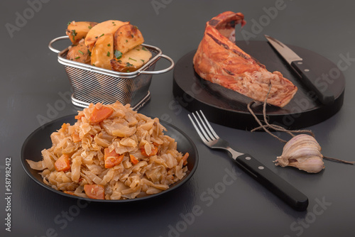 Stewed cabbage - bigos in a black plate on the background of a piece of smoked meat and baked potatoes. Country style breakfast photo