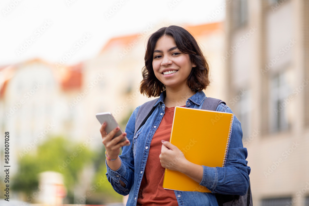 Outdoor Portrait Of Smiling Arab Female Student With Smartphone And Workbooks Outdoor Portrait Of Smiling Arab Female Student With Smartphone And Workbooks