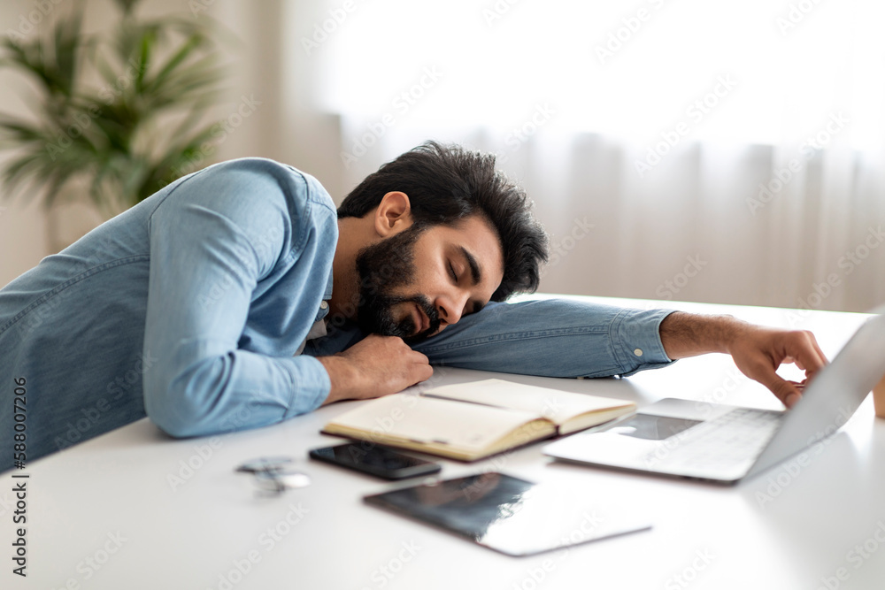 Tired indian male freelancer sleeping at desk in home office Tired indian male freelancer sleeping at desk in home office