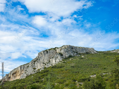 Wild landscape of the Alpilles in Provence in France where a rocky outcrop rises from a hill covered in scrubland under a beautiful blue sky adorned with pretty light fluffy white clouds