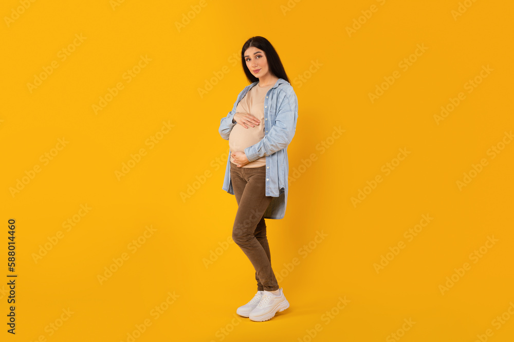 Full length portrait of beautiful pregnant lady embracing belly and looking at camera, yellow background, free space Full length portrait of beautiful pregnant lady embracing belly and looking at camera, yellow background, free space