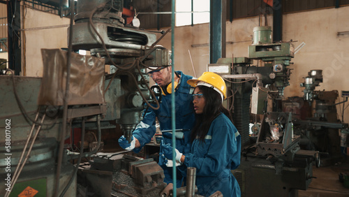 African factory worker in safety wear is checking a piece of industrial metal while Caucasian coworker help her to combine metal part to machine. Two production engineers are working in metal factory.